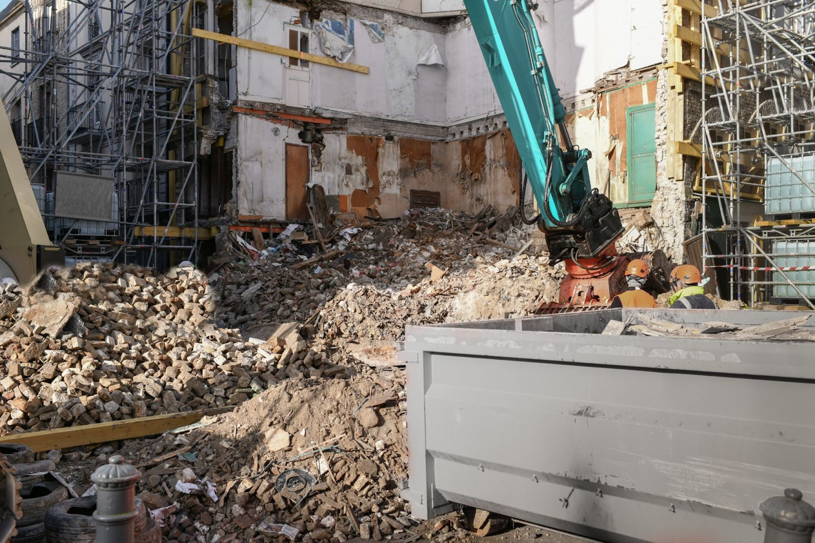 An excavator tears down a building while a gray dumpster is on standby to collect the debris.