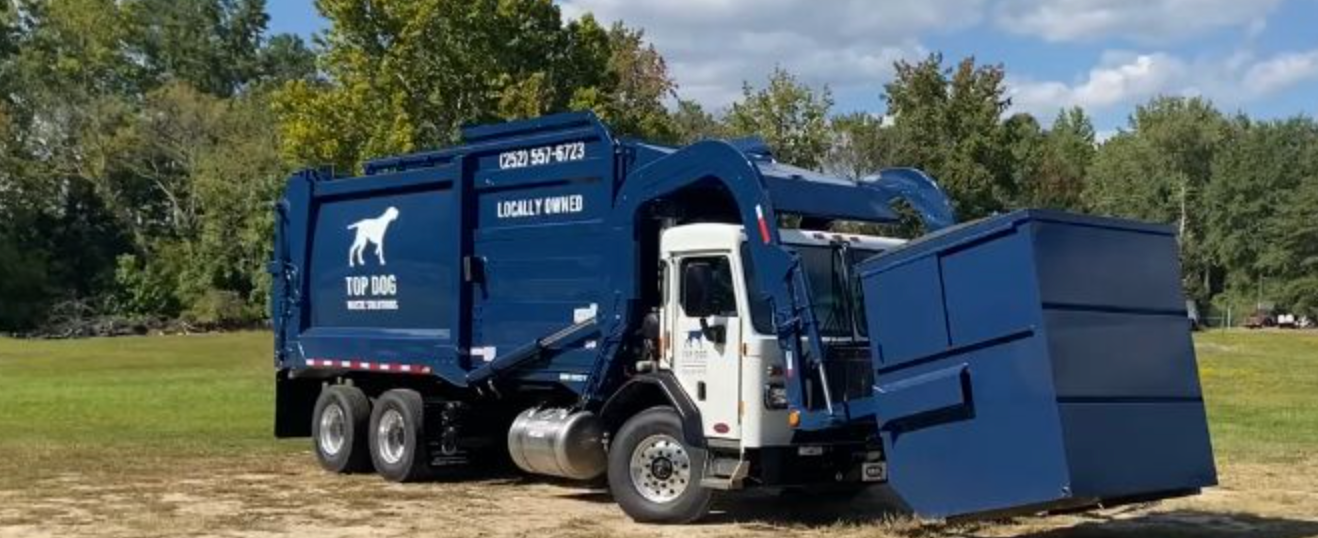 Trash truck lifting a dumpster on the grass with trees in the background. 